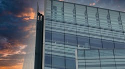 A modern glass and steel building against a colorful sunset sky. The building s facade features large windows and a grid pattern, while the sky is a blend of orange, purple, and blue